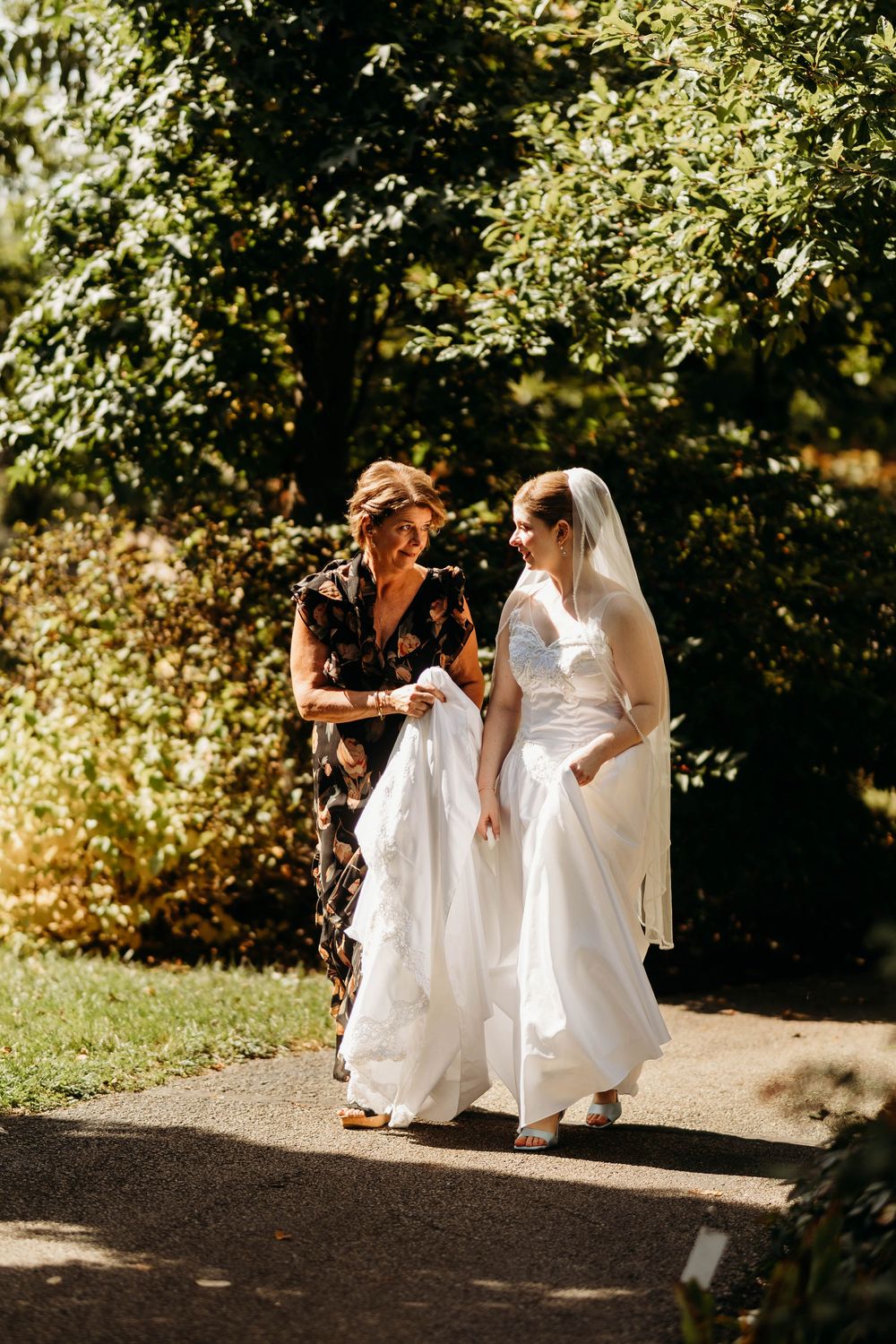 A bride in a white lace gown and veil walks with an older woman through a sunlit garden path.