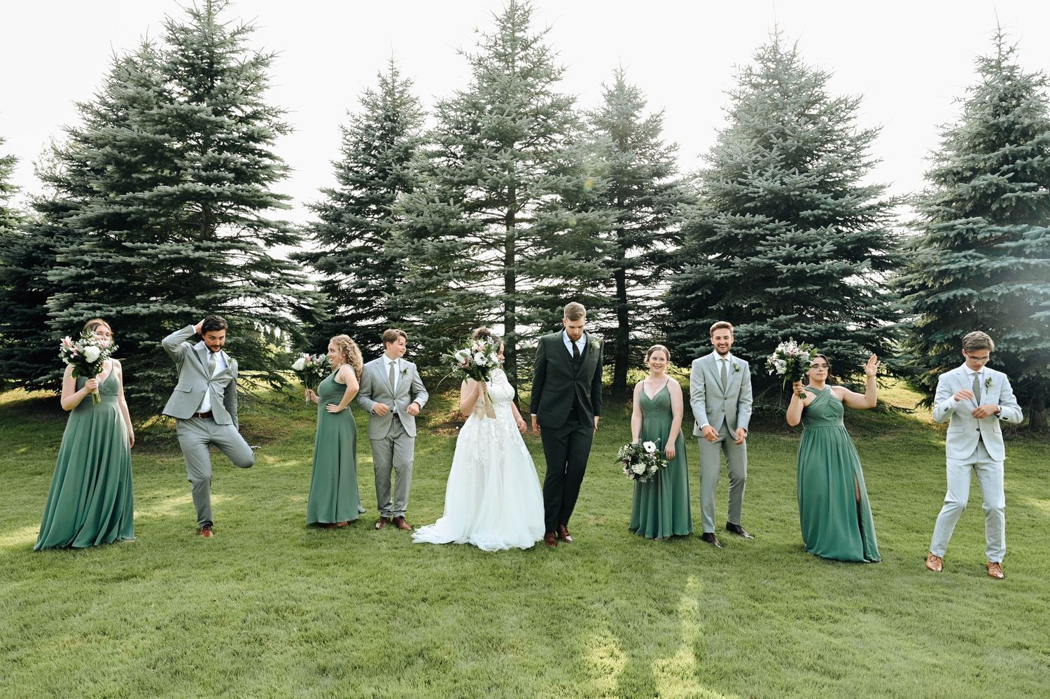 Wedding party poses playfully against row of evergreen trees in coordinated attire.