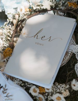 A white vow book with gold 'her' text surrounded by delicate dried flowers.