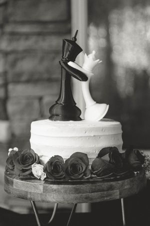 A wedding cake topped with black and white chess piece figurines surrounded by dark and light roses.