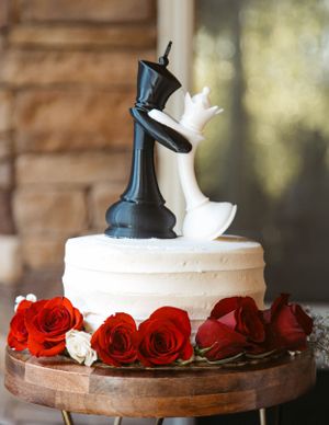Chess-themed wedding cake topped with black king and white queen pieces, decorated with red and white roses on wooden stand.