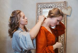 Two friends share a lighthearted moment while getting ready in front of an ornate mirror, one wearing orange and one in blue.