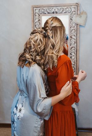 Two women in blue and rust colored dresses embrace while looking in an ornate vintage mirror.