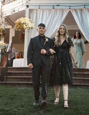 Two people in formal attire walk together on grass in front of a white draped gazebo decorated with yellow flowers.
