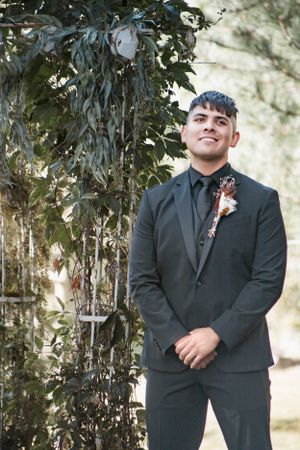 A person in a dark formal suit and boutonniere stands beside leafy foliage at an outdoor event.