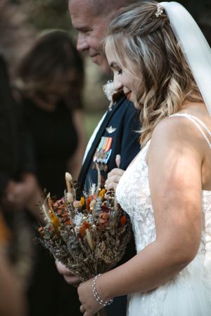A bride in a white dress holds a rustic dried flower bouquet while standing next to someone in military dress uniform.