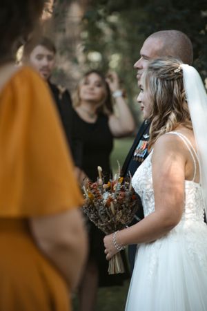 A bride in a white dress holds a rustic bouquet while standing with guests at an outdoor wedding ceremony.