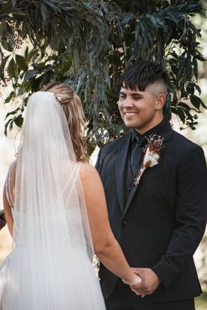A wedding ceremony under trees with the couple holding hands while exchanging vows.