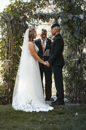 A wedding ceremony takes place under a lush greenery arch with an officiant presiding over the couple exchanging vows.