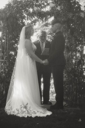 A black and white wedding ceremony photo shows a couple exchanging vows under a leafy garden arch with an officiant.