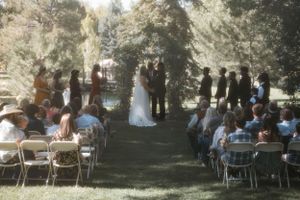 An outdoor wedding ceremony takes place in a wooded setting with guests seated on white folding chairs.