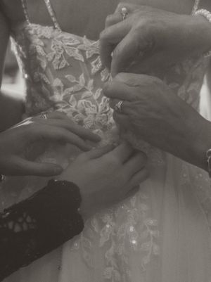 Multiple hands carefully adjusting delicate lace details on a white wedding dress in a black and white photograph.