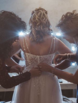 Bridesmaids helping to button up the back of a white wedding dress in dramatic lighting.