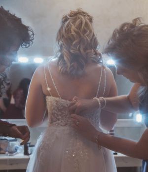 Bridesmaids helping adjust a wedding dress during preparation with vanity lights in the background.