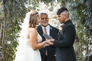 A couple exchanges wedding vows during an outdoor ceremony surrounded by greenery.