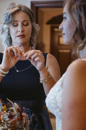 A woman in a black dress helps adjust jewelry during wedding preparations.