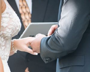 Close-up view of hands exchanging rings during an elegant wedding ceremony with lace dress and gray suit details.