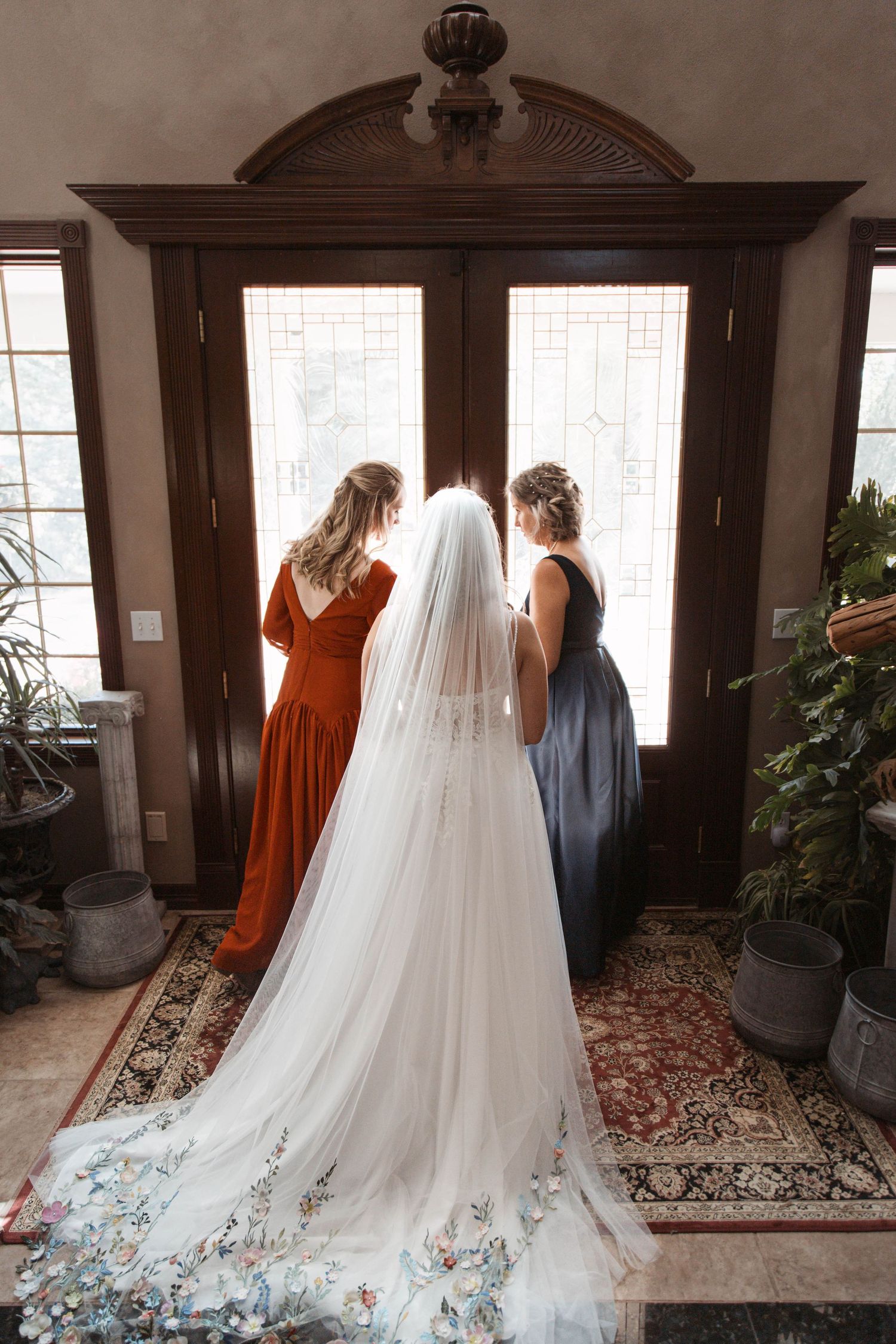 A wedding dress with a flowing floral-embellished train is photographed from behind near an elegant wooden doorway.