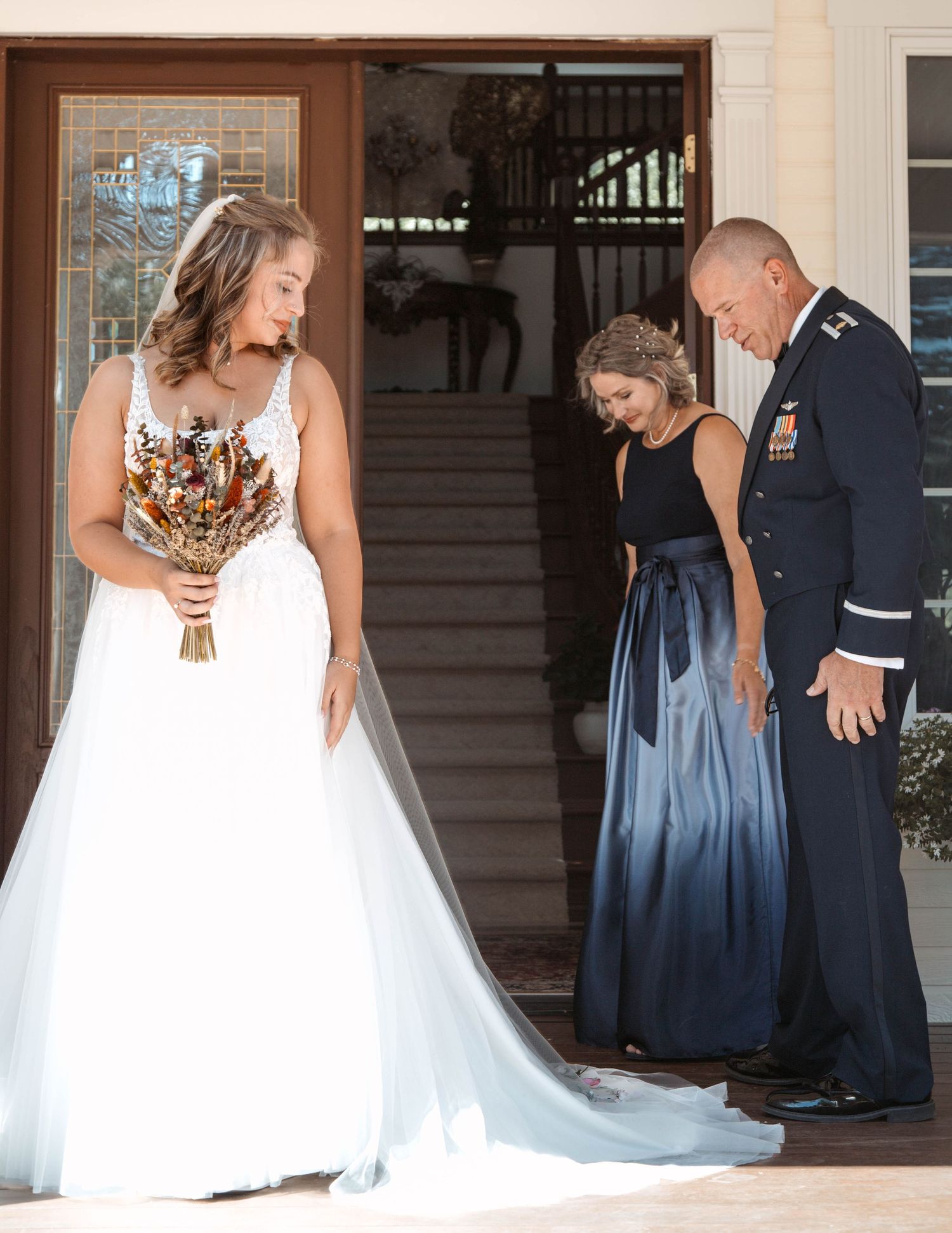 A bride in a white gown stands with two guests in formal attire on the steps of an elegant building entrance.