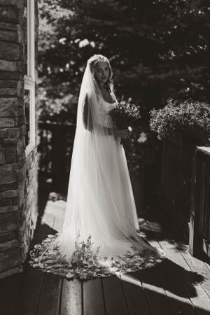 A bride in a flowing white gown and veil stands on a rustic wooden porch in dramatic black and white lighting.