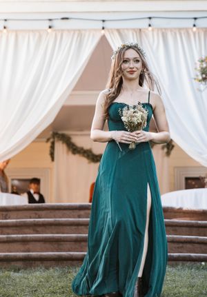 Woman in an elegant emerald green formal dress with slit holding a small bouquet under a white draped canopy.