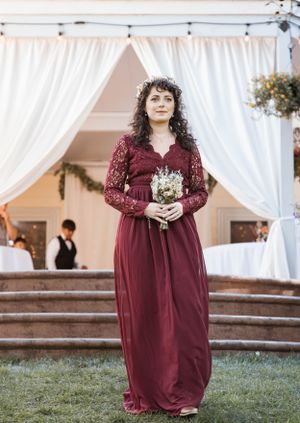 A person in a burgundy lace bridesmaid dress holds a bouquet under a white draped wedding arch.