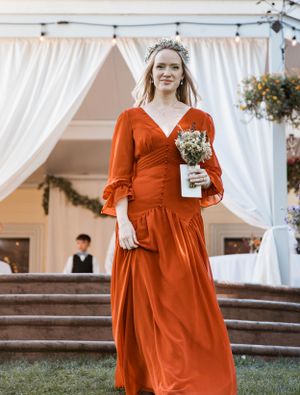 A person in a burnt orange bridesmaid dress holds a small bouquet under a white tent at an outdoor wedding.