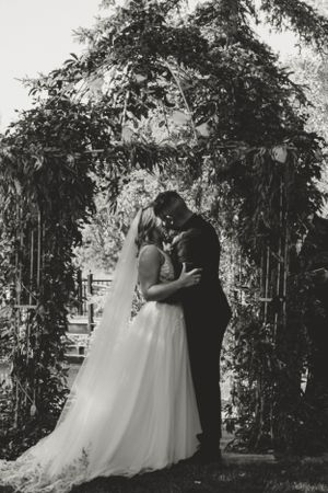 A romantic black and white wedding photo of a couple kissing under a floral arch in an outdoor garden setting.