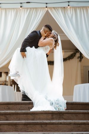 A romantic wedding photo of a couple sharing a dramatic kiss on stone steps with flowing white drapery in the background.