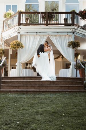 A romantic wedding couple embraces on wooden steps under an elegant white draped gazebo with a balcony above.