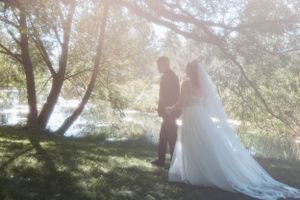 A couple in wedding attire stands together in a dreamy sunlit garden setting by a lake.