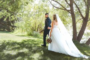 A couple in wedding attire poses together under a tree in a sunny garden setting.