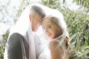 A romantic wedding couple shares an intimate moment under a veil with sunlight streaming through green foliage.