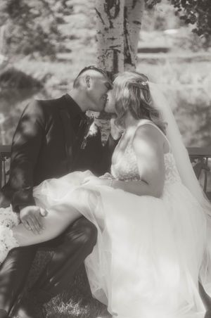 A romantic black and white wedding photo of a couple sharing an intimate moment on a bench outdoors.