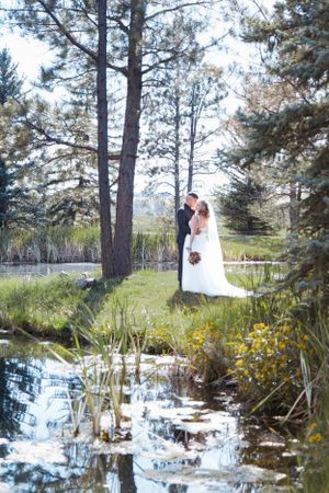 A romantic wedding couple embraces by a reflective pond surrounded by tall pine trees in a natural outdoor setting.
