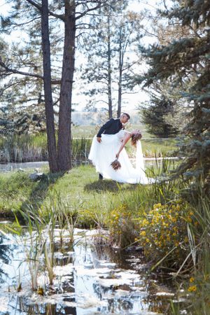 A romantic wedding scene by a pond surrounded by tall pine trees and yellow wildflowers in a natural outdoor setting.