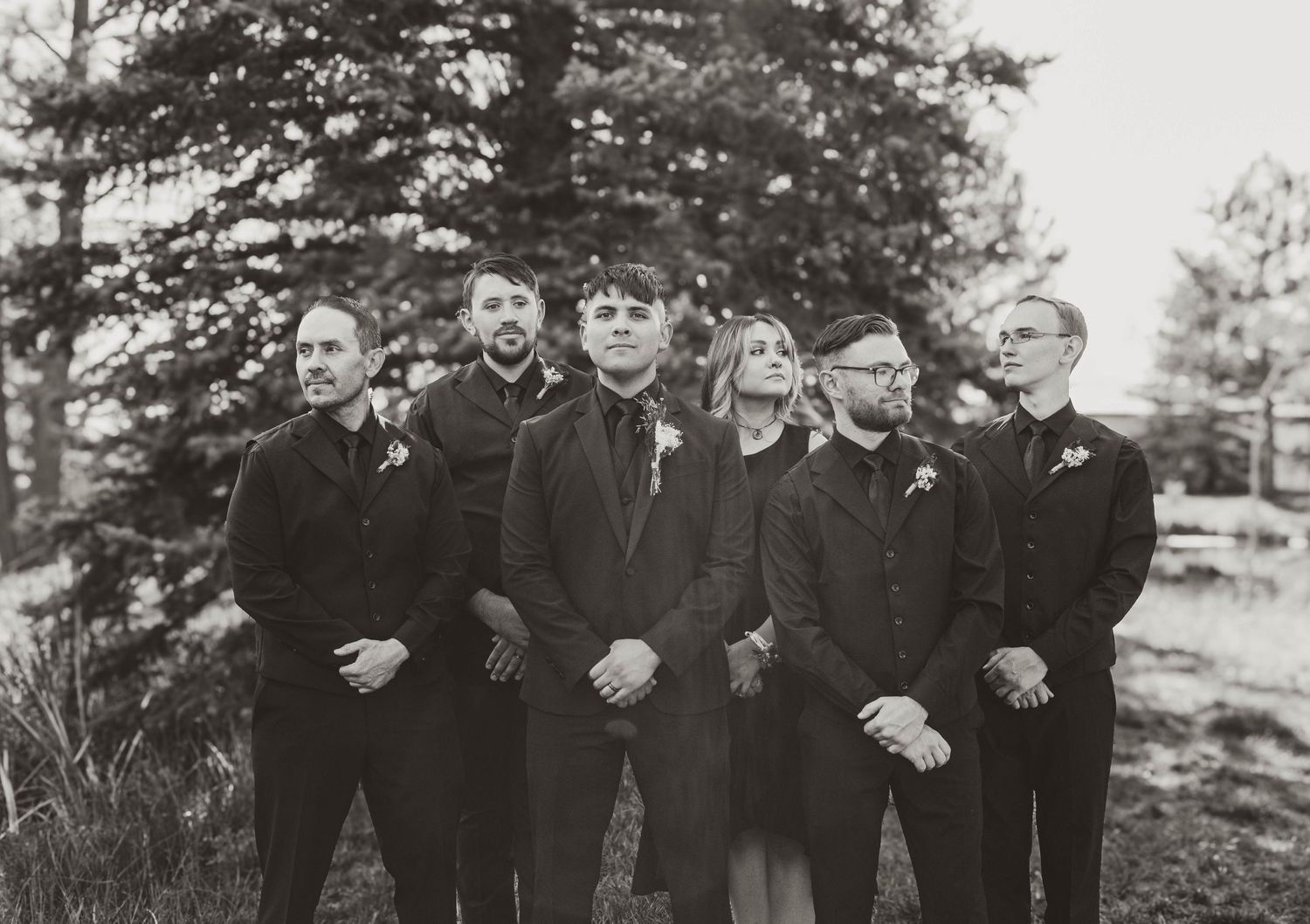 A black and white photo of a wedding party in formal attire standing together outdoors with pine trees in the background.