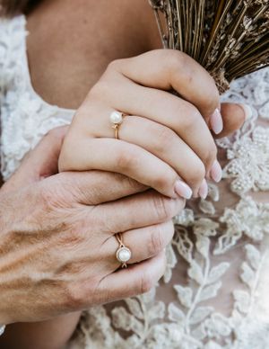 Close-up of hands with engagement rings holding dried wheat against lace fabric.