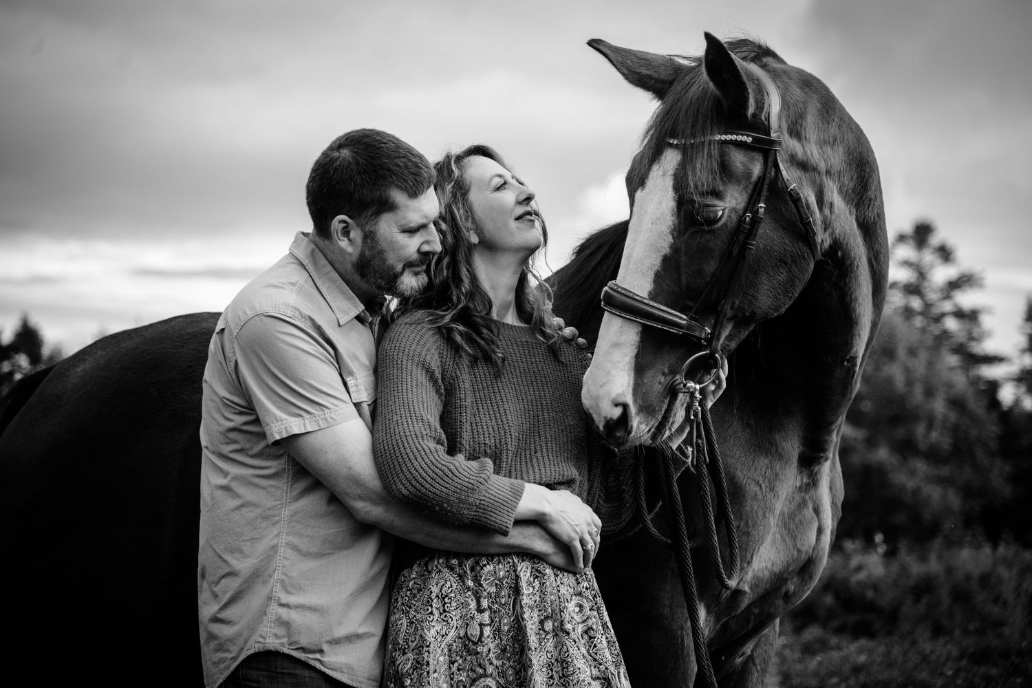 Black and white photo of a couple embracing beside a dark horse under a cloudy sky. Photo by Xenophon Photography in Maine.