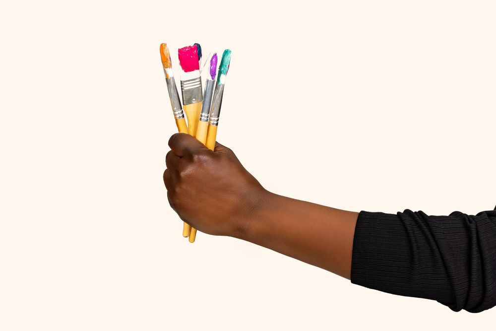Close-up of a dark-skinned hand holding multiple colorful paint brushes against a light beige background.