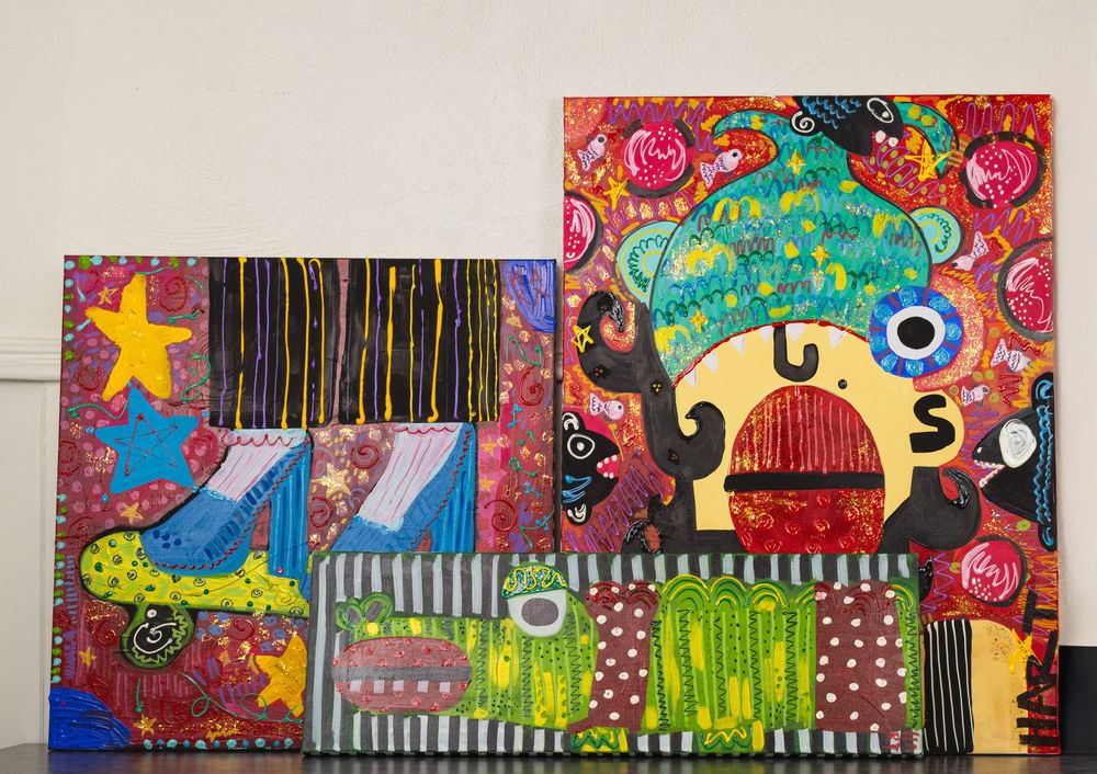 Two colorful folk art paintings displayed together featuring whimsical characters, bold patterns, stars, and bright mixed colors.