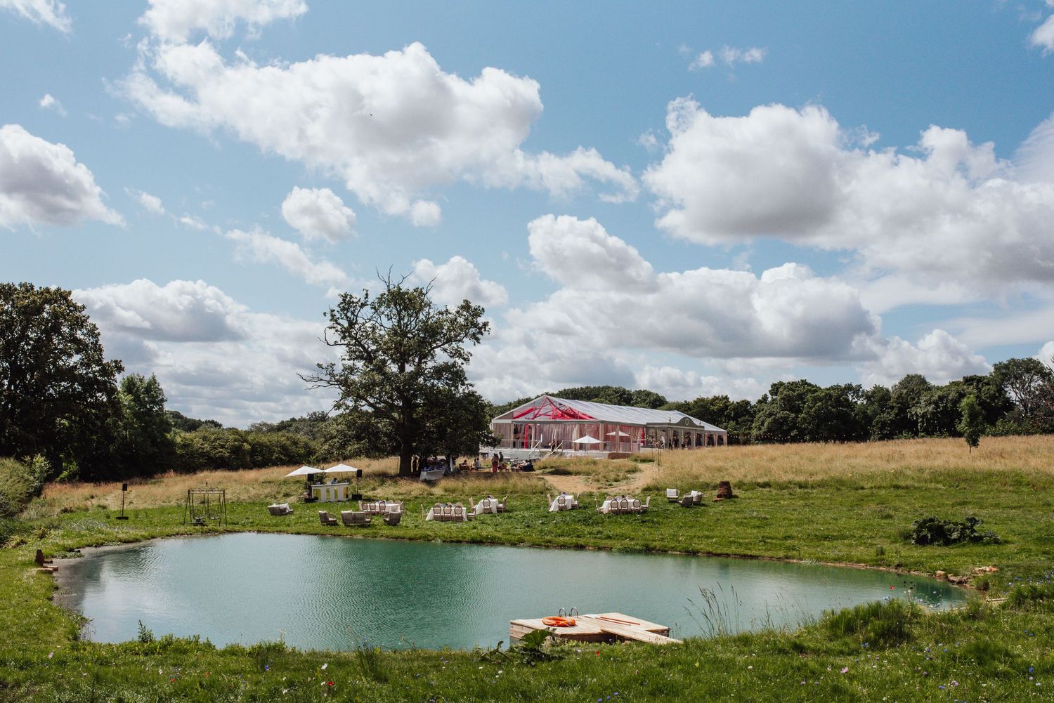 A scenic farm pond surrounded by green pastures with a red barn and fluffy white clouds in the blue sky.