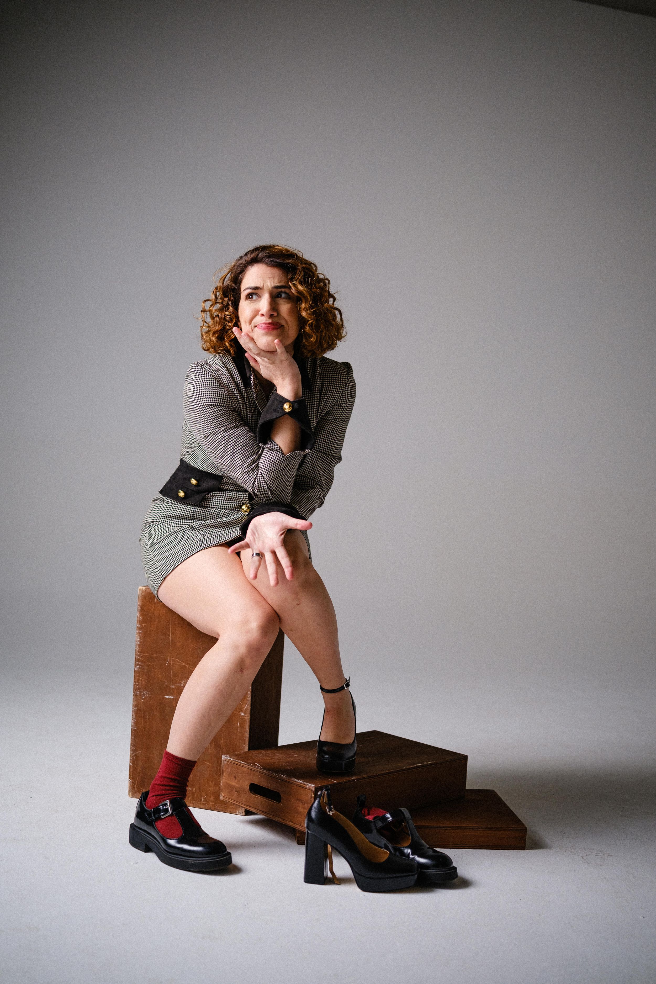 Model poses playfully on wooden box wearing gray blazer and black shoes with red socks in studio setting.