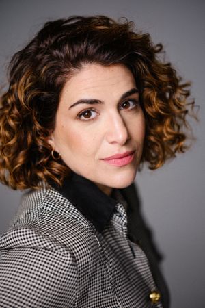 Professional studio portrait showing curly brown hair and checkered blazer against dark background.