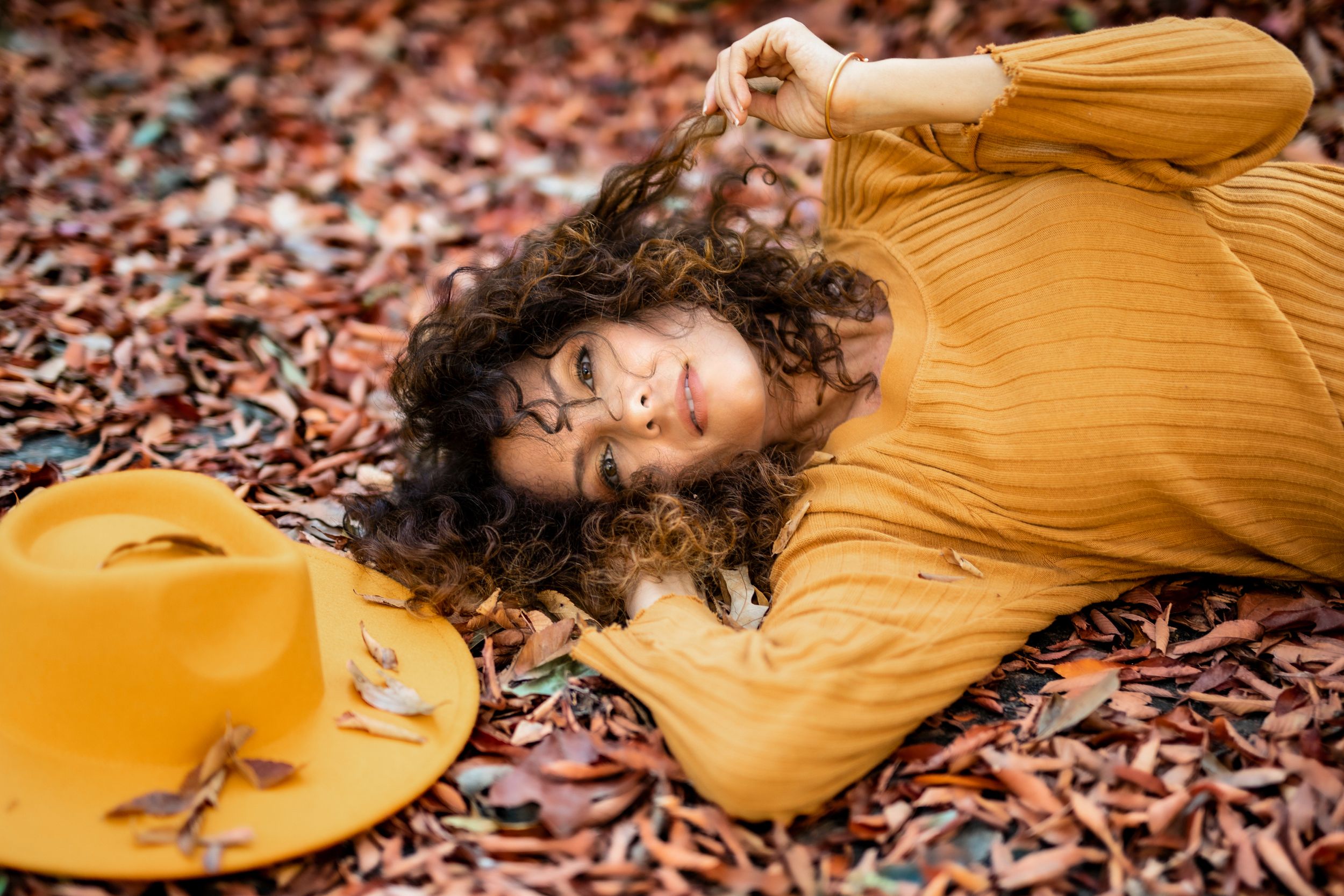 Woman in yellow dress laying in fall leaves twirling her hair – fantasy creative portrait photography in Windsor, Ontario.
