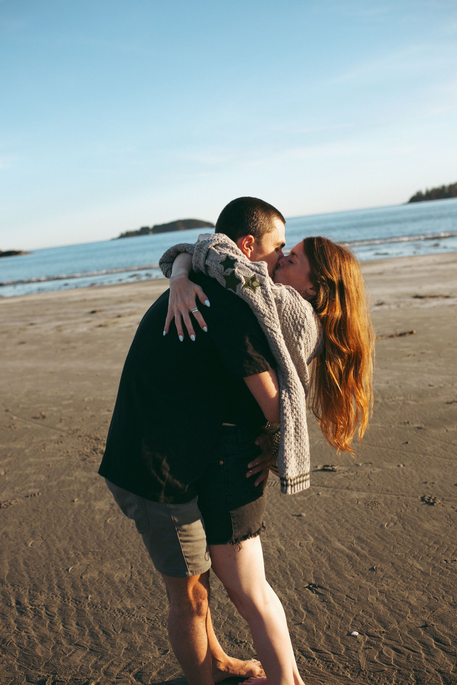 Couple sharing a romantic kiss on a sandy beach at sunset, with ocean waves and blue sky in the background.