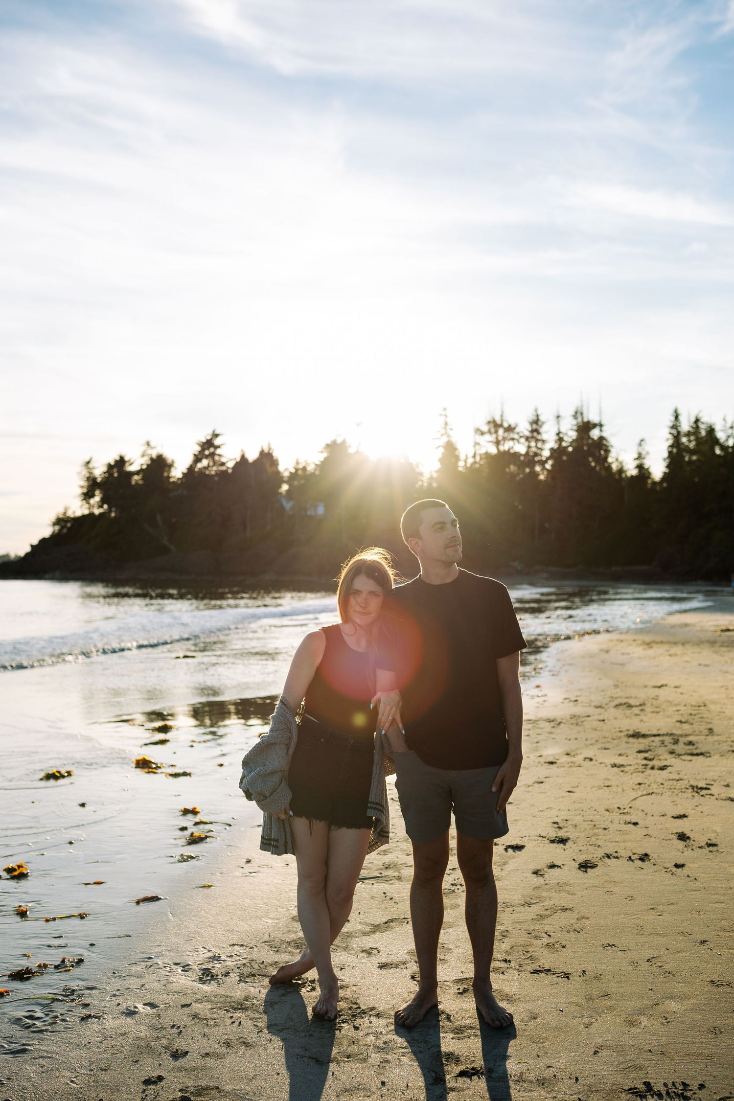 Couple standing together on a sunlit beach with lens flare, ocean waves, and tree-lined shore in the background.