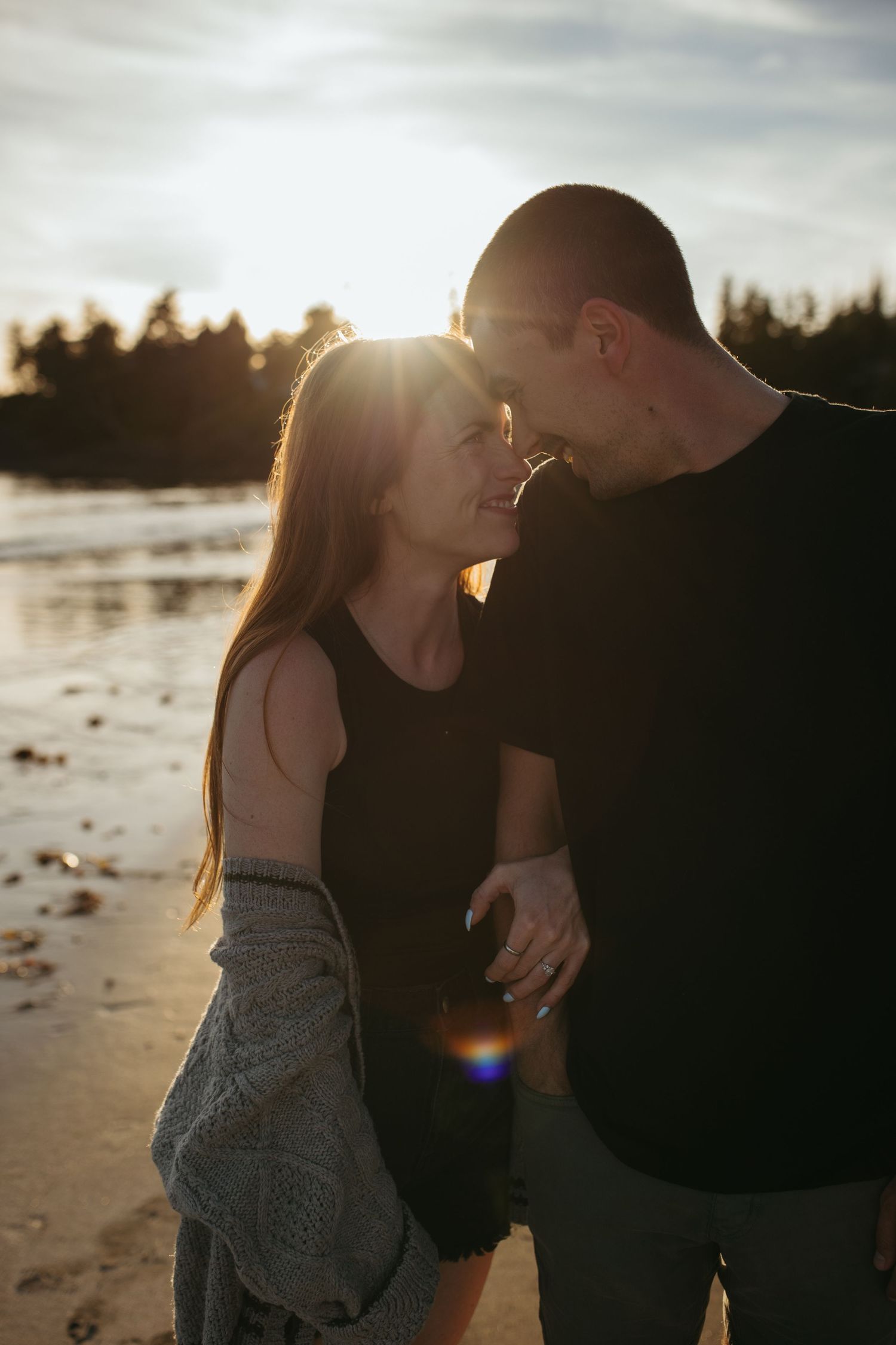 Couple sharing a romantic moment on a beach at golden sunset, backlit by warm sunlight with trees in background.