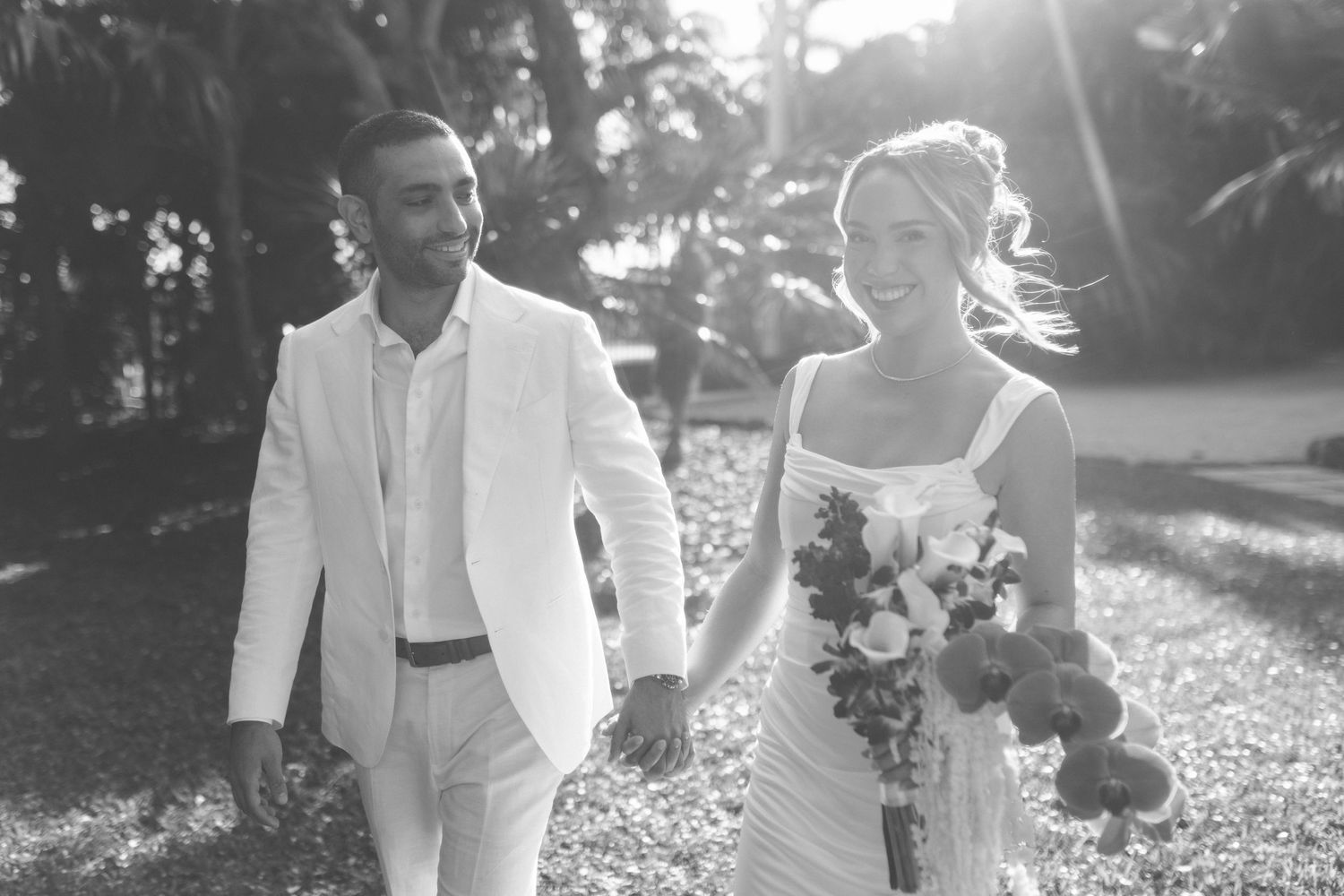 A couple walks together in sunlit garden wearing elegant white wedding attire.