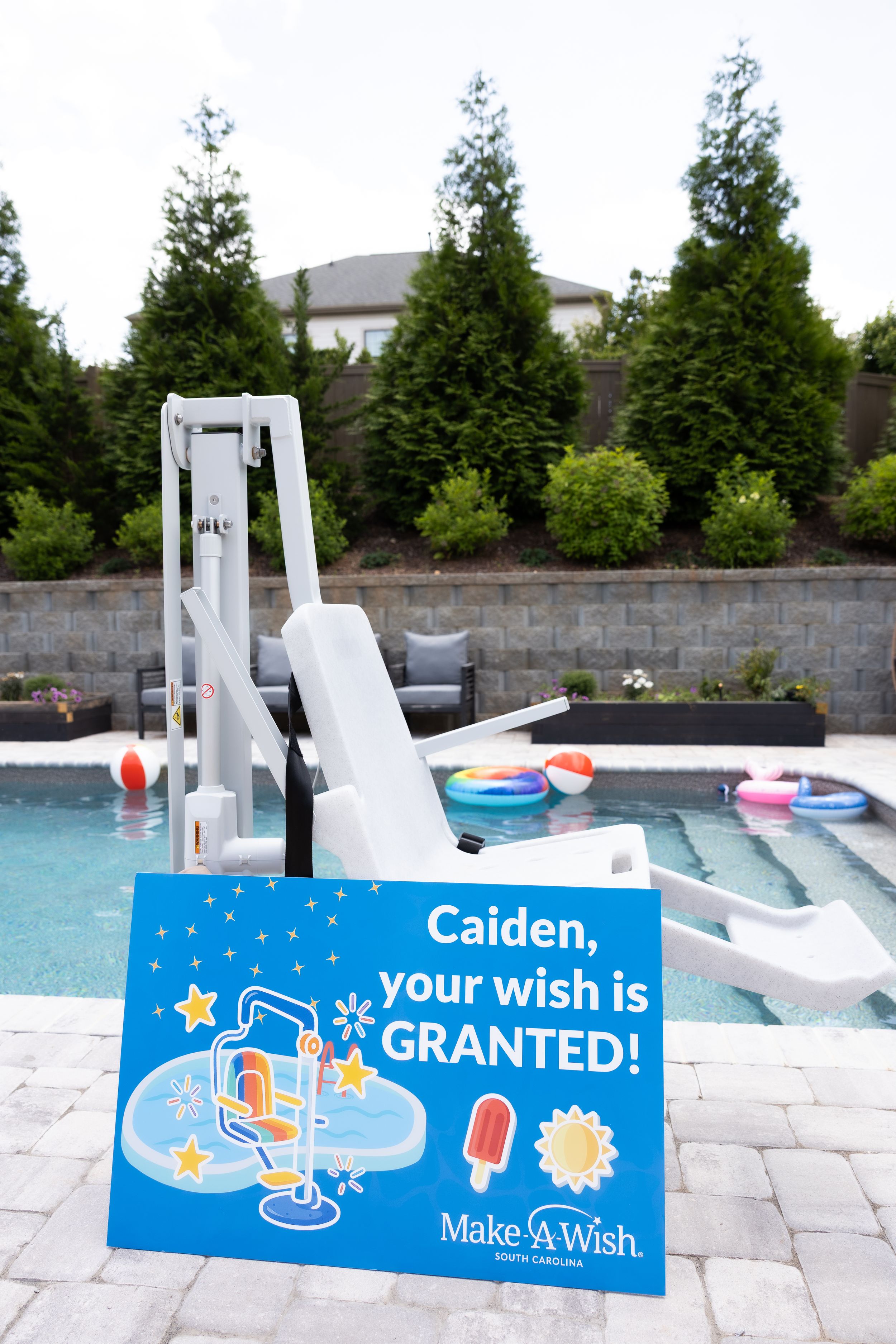 A Make-A-Wish Foundation sign and pool lift chair visible beside an outdoor swimming pool with inflatable toys.